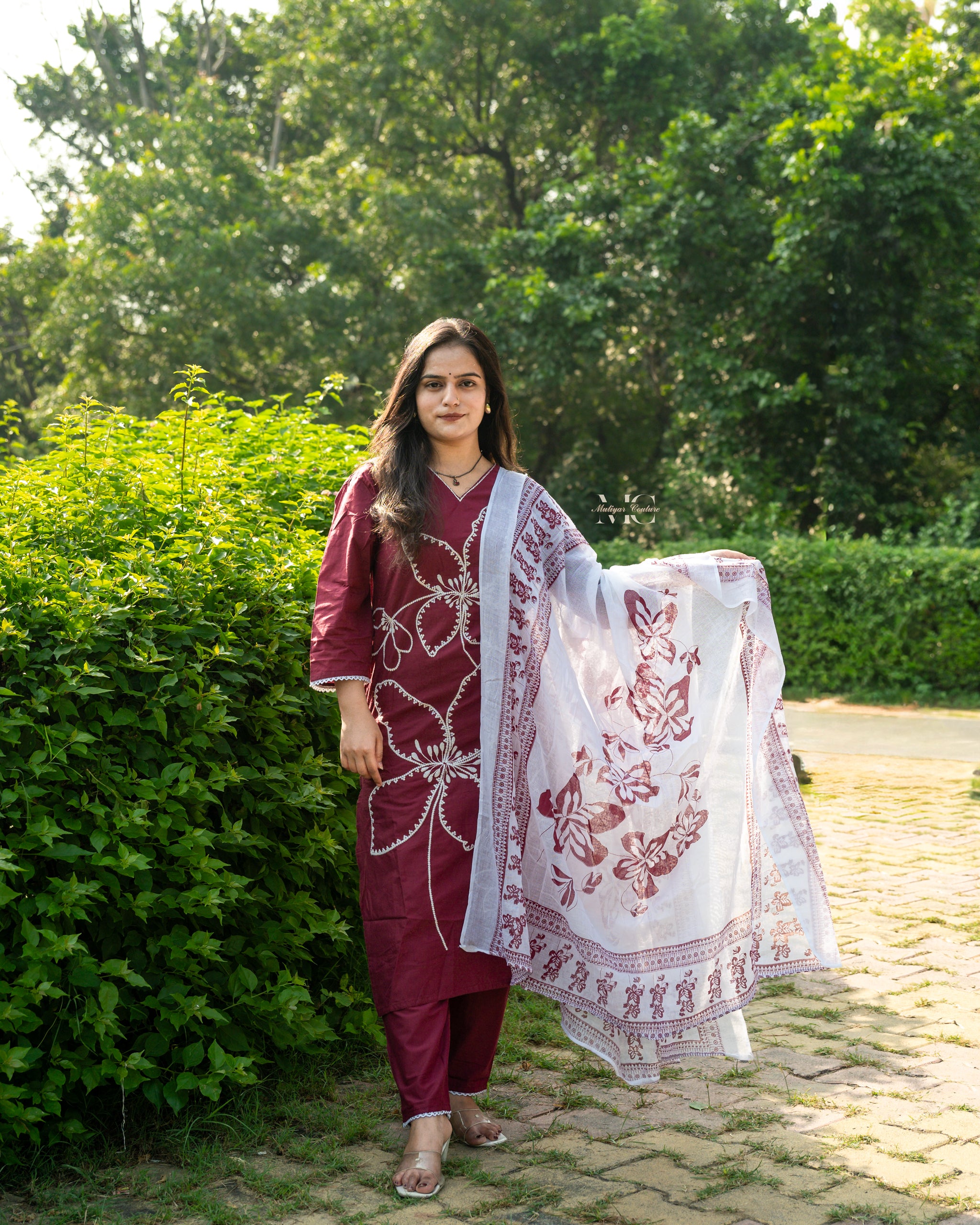 Woman in traditional maroon and white outfit standing outdoors with greenery in the background